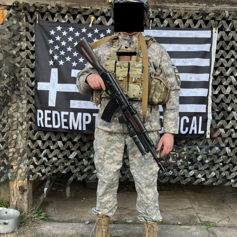 Person in military uniform holding a rifle in front of flags with text