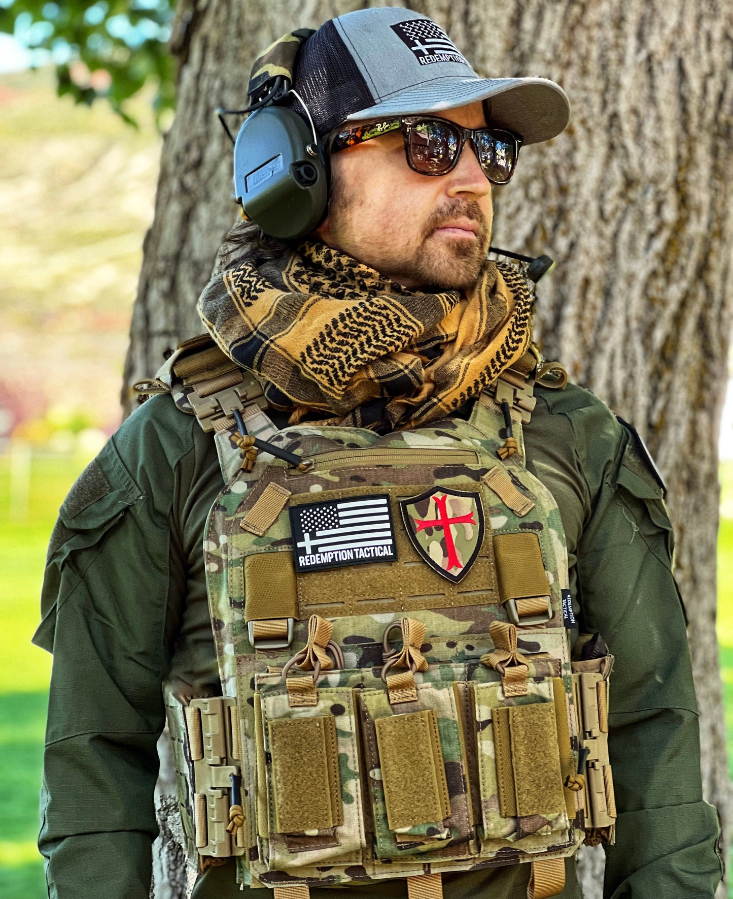 Person wearing tactical gear with a camouflage vest and American flag patches outdoors.
